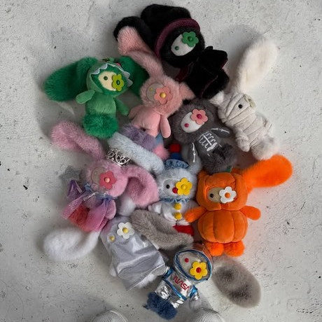 A pile of assorted plush toys and small stuffed characters arranged on a light concrete floor, with a person's white sneakers visible at the bottom edge.